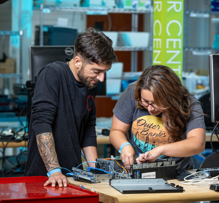 A male IT Academy student (left) and a female IT Academy student (right) hover over an opened PC. The female student has a small tool in her hand and is working inside of the computer.